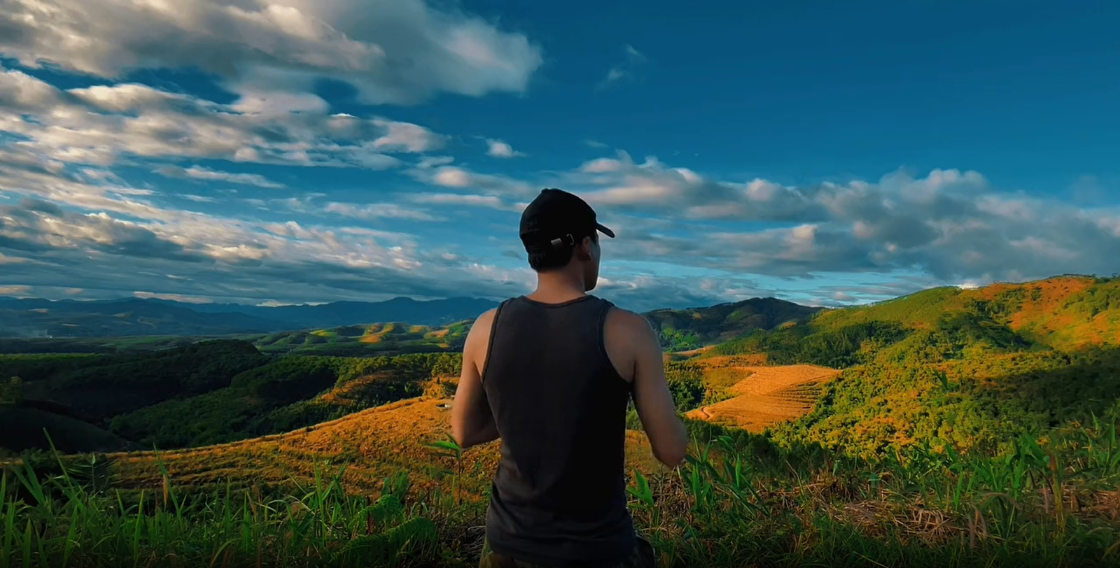 Colour-graded frame of a man in a cap looking out over sunlit green hills under a wide cloudy sky.
