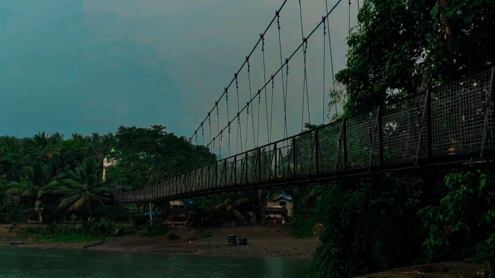 Teal-graded dusk frame of a suspension footbridge crossing a river, with palms and houses along the far bank.