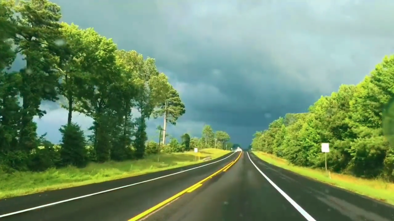 Driving view down a two-lane highway lined with bright green trees, a dark storm sky building ahead.